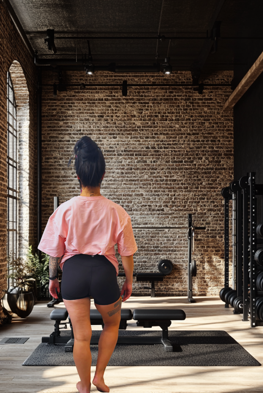 Back view of Women's Pink Oversized Tee - BulkBully's G.O.A.L.S Collection, styled casually in a fitness studio with brick walls and gym equipment.