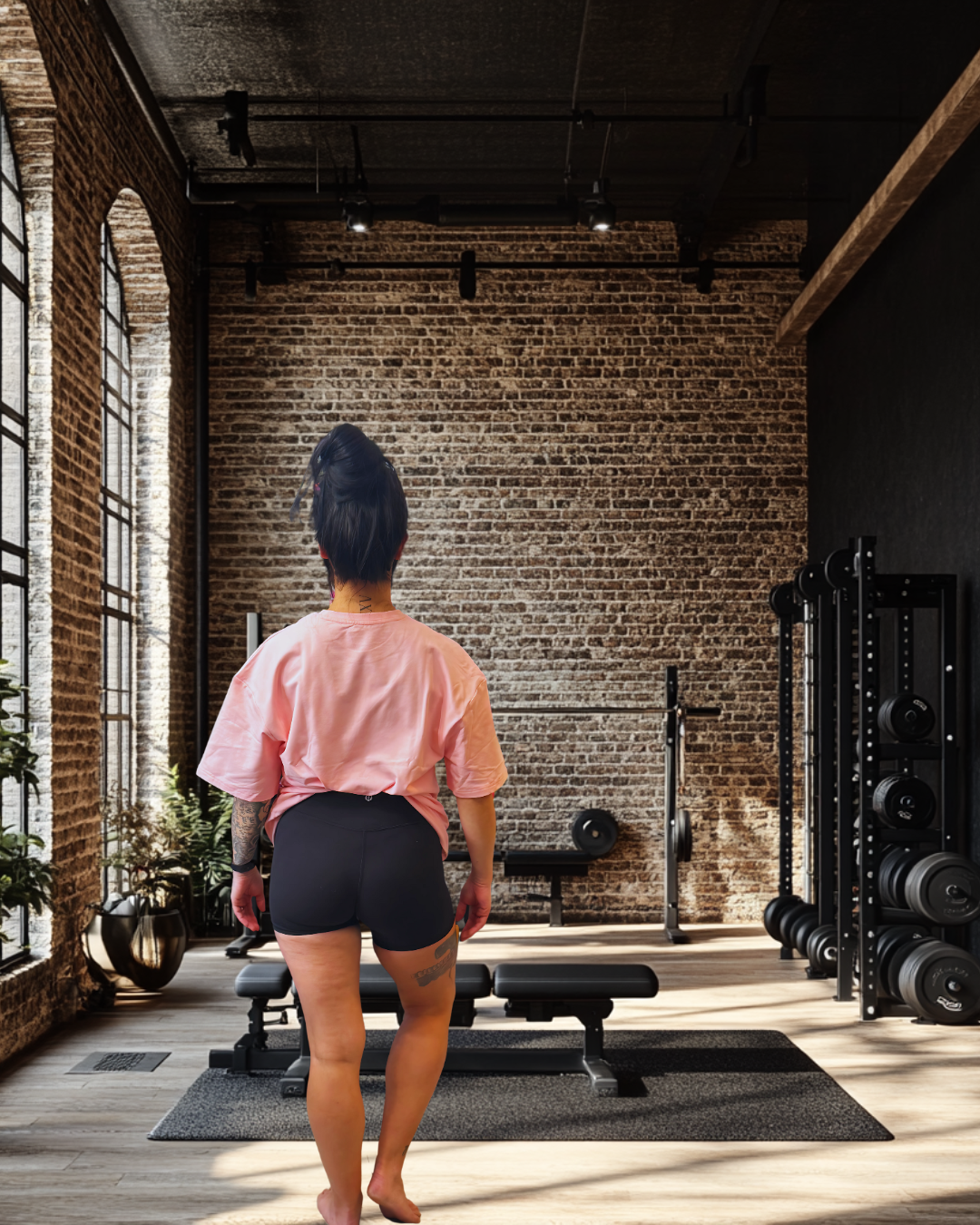 Back view of Women's Pink Oversized Tee - BulkBully's G.O.A.L.S Collection, styled casually in a fitness studio with brick walls and gym equipment.